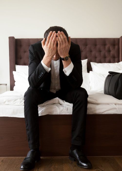 Upset stressed man in business suit sitting with head in hands on bed edge beside  handbag with belongings. Leaving home because of divorce, problems in relations or business, loss through loan debts